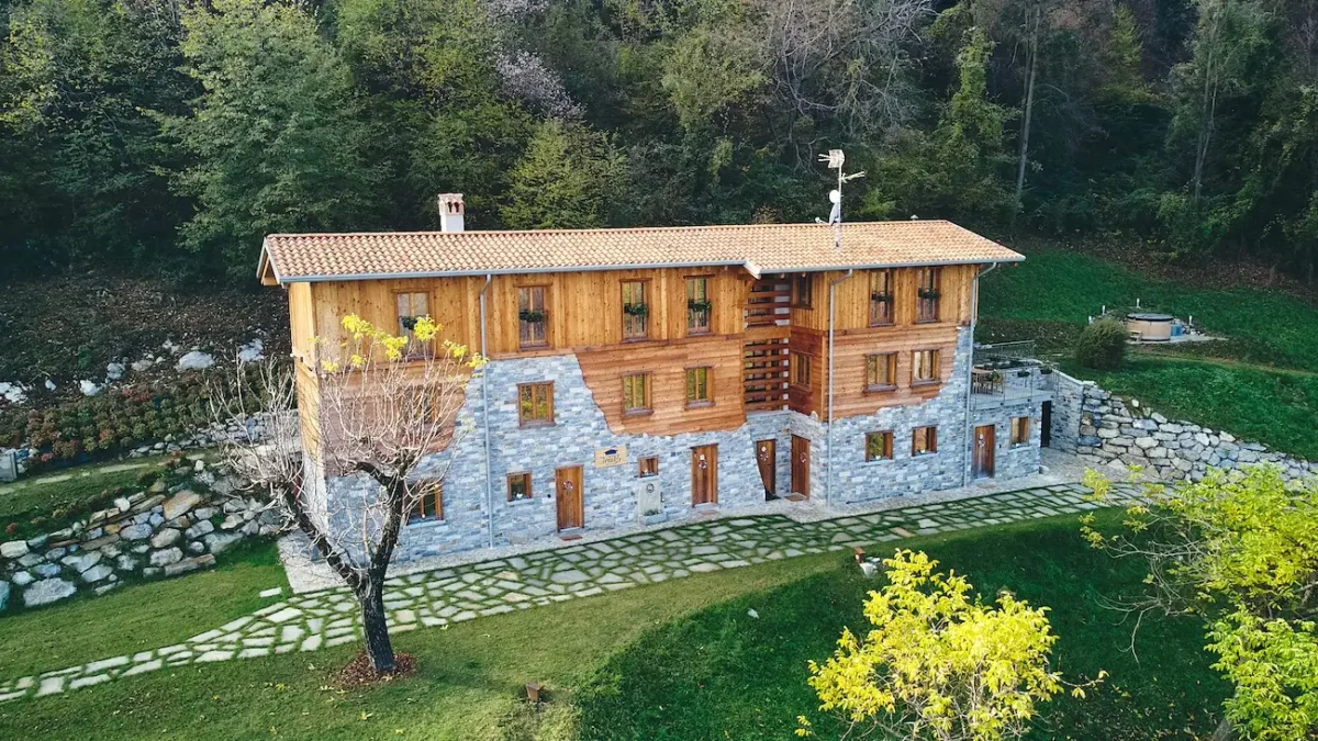 Rustic holiday apartment building in Bellagio, Lake Como, featuring a mix of stone and wood exterior, surrounded by trees and greenery, with a hot tub area visible in the garden.